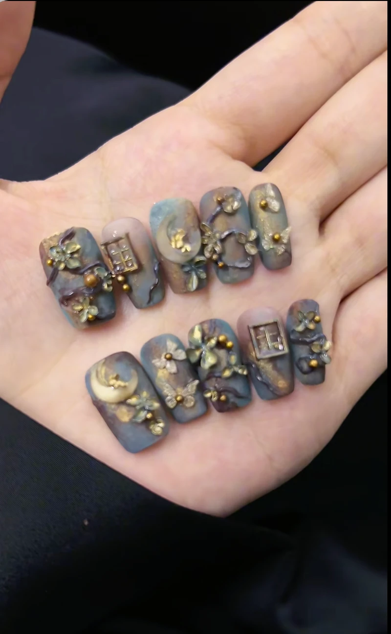 Ornate gray-blue and purple press-on nails with gold botanical and moon details, held in a palm